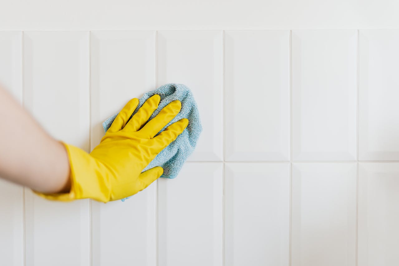 A hand in a yellow glove wiping a tiled wall with a cloth, demonstrating cleanliness.