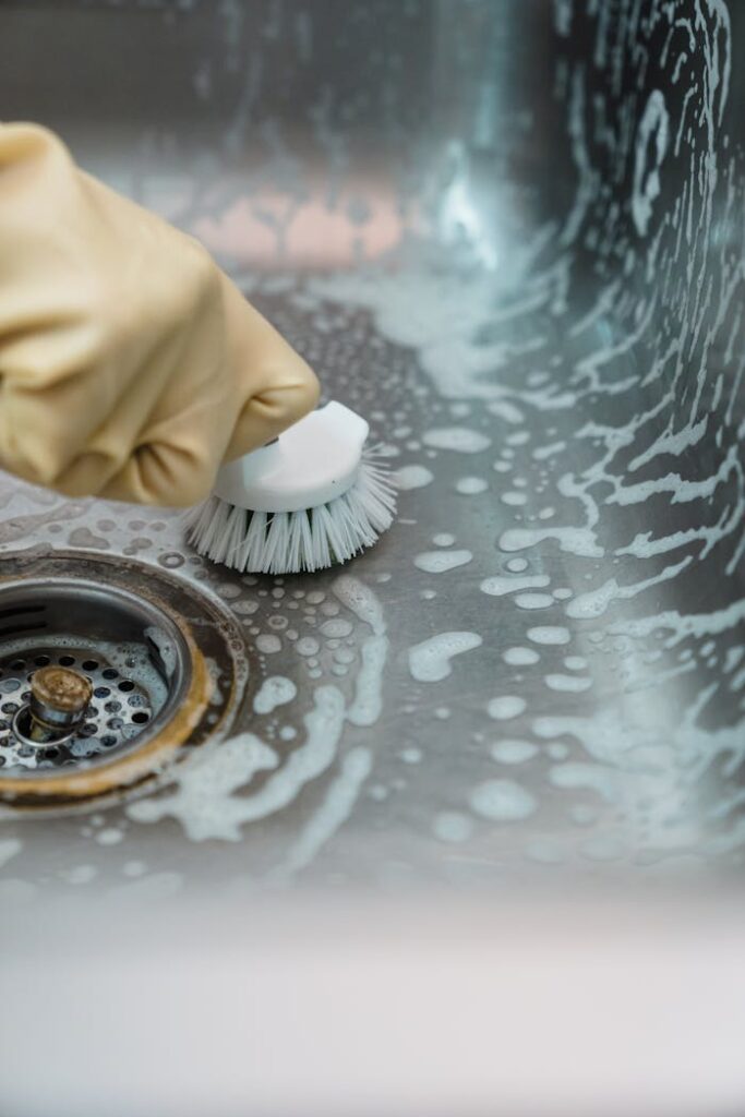 Close-up of a hand in gloves scrubbing a soapy kitchen sink with a brush.